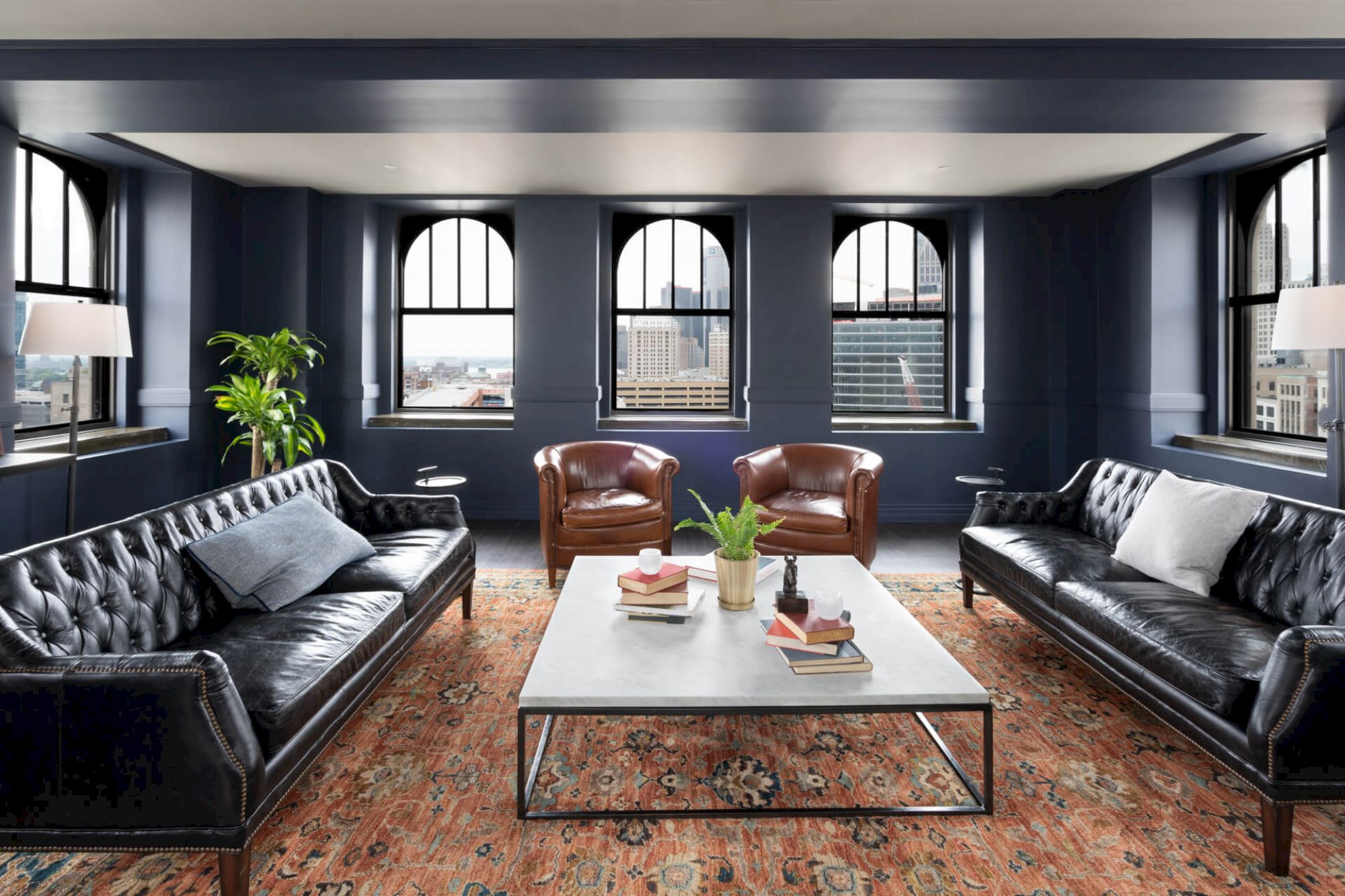 The image shows a stylish living room with dark sofas, leather chairs, a coffee table, large windows, and a patterned rug, exuding elegance.