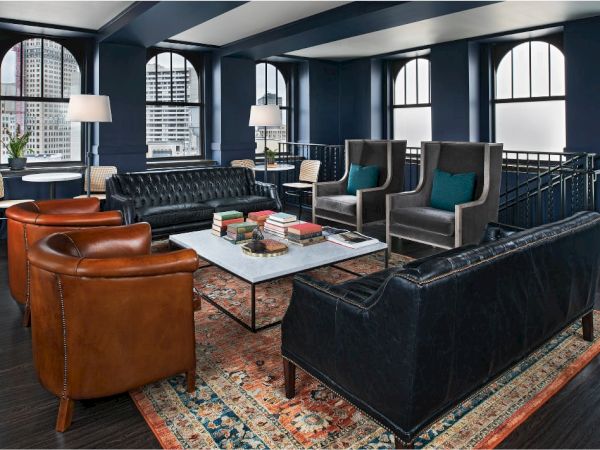 A stylish lounge with leather sofas, armchairs, books on a marble table, floor lamps, and an intricate rug, set in a room with arched windows.
