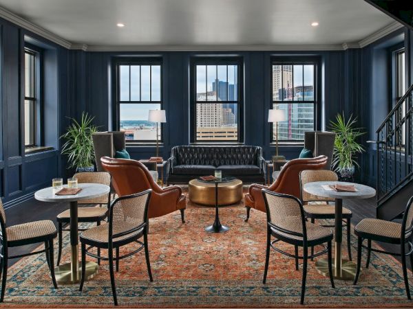 A stylish lounge with leather chairs, a black sofa, marble tables, and large windows overlooking a cityscape, adorned with plants and lamps.