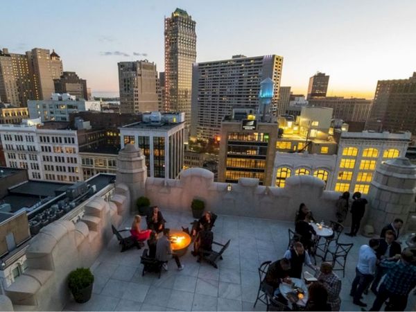 Rooftop gathering at sunset with people around tables and a fire pit, overlooking a city skyline with tall buildings and evening lights.