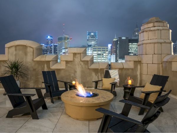 A rooftop patio with a fire pit, surrounded by chairs, and a city skyline in the background at night.