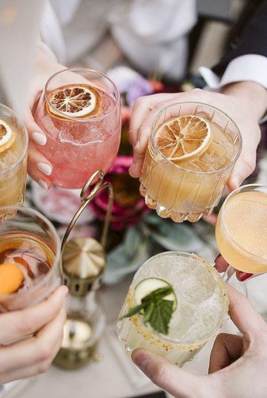 A group of people raising colorful cocktails garnished with citrus and herbs, suggesting a toast or celebration.