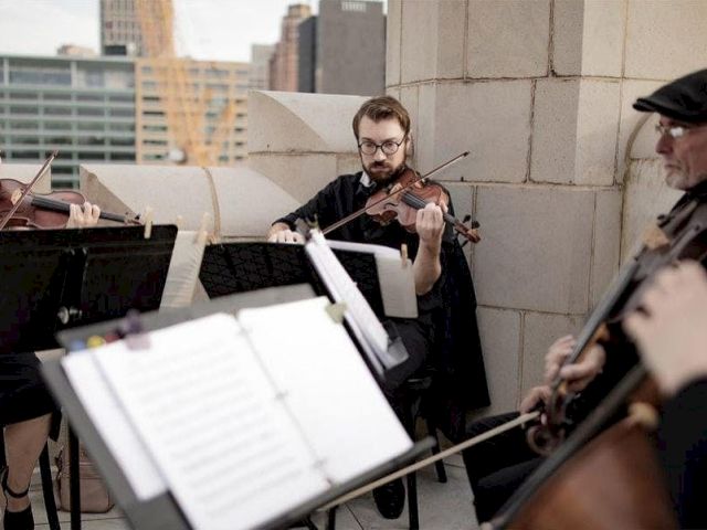 A string quartet is playing music outdoors on a rooftop, with city buildings in the background.