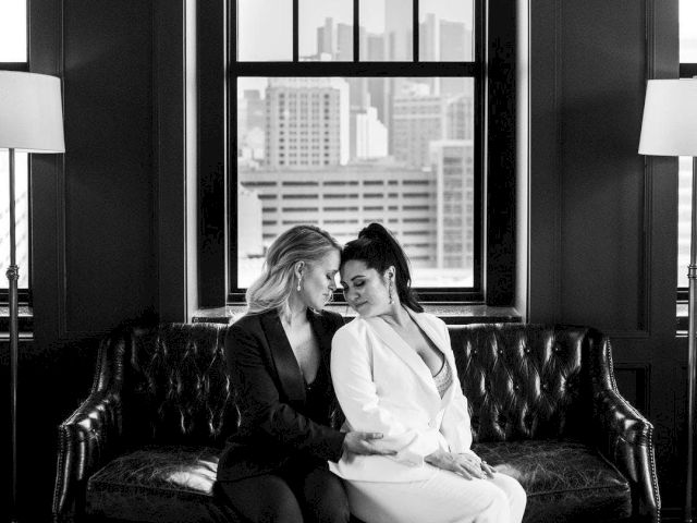 A black-and-white image of two people in suits sitting closely on a leather couch in front of large windows with a cityscape view.