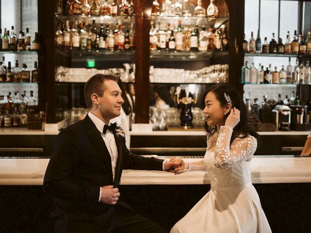 A couple in formal attire shares a moment at a bar, surrounded by shelves of bottles and glasses, capturing a wedding or special occasion.