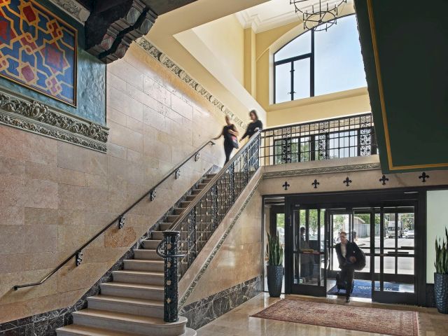 The image shows a spacious, elegant lobby with a staircase, decorative tiles, and people entering through glass doors.