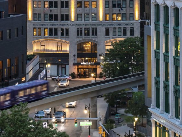 An elevated train moves through a city scene at dusk, with cars below and lit buildings surrounding the area, creating a lively urban atmosphere.