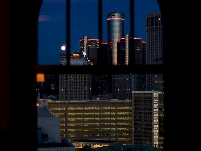 A cityscape at night viewed through a window, with tall buildings and a lit tower under a dark blue sky.