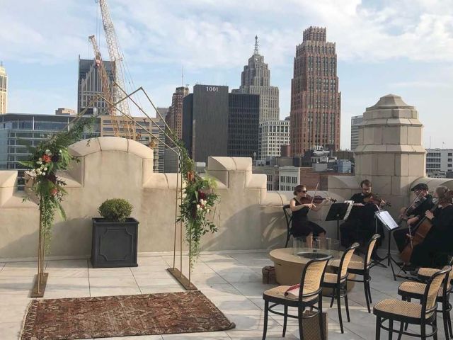Rooftop setup with chairs and floral decorations facing city skyscrapers; string musicians perform nearby.