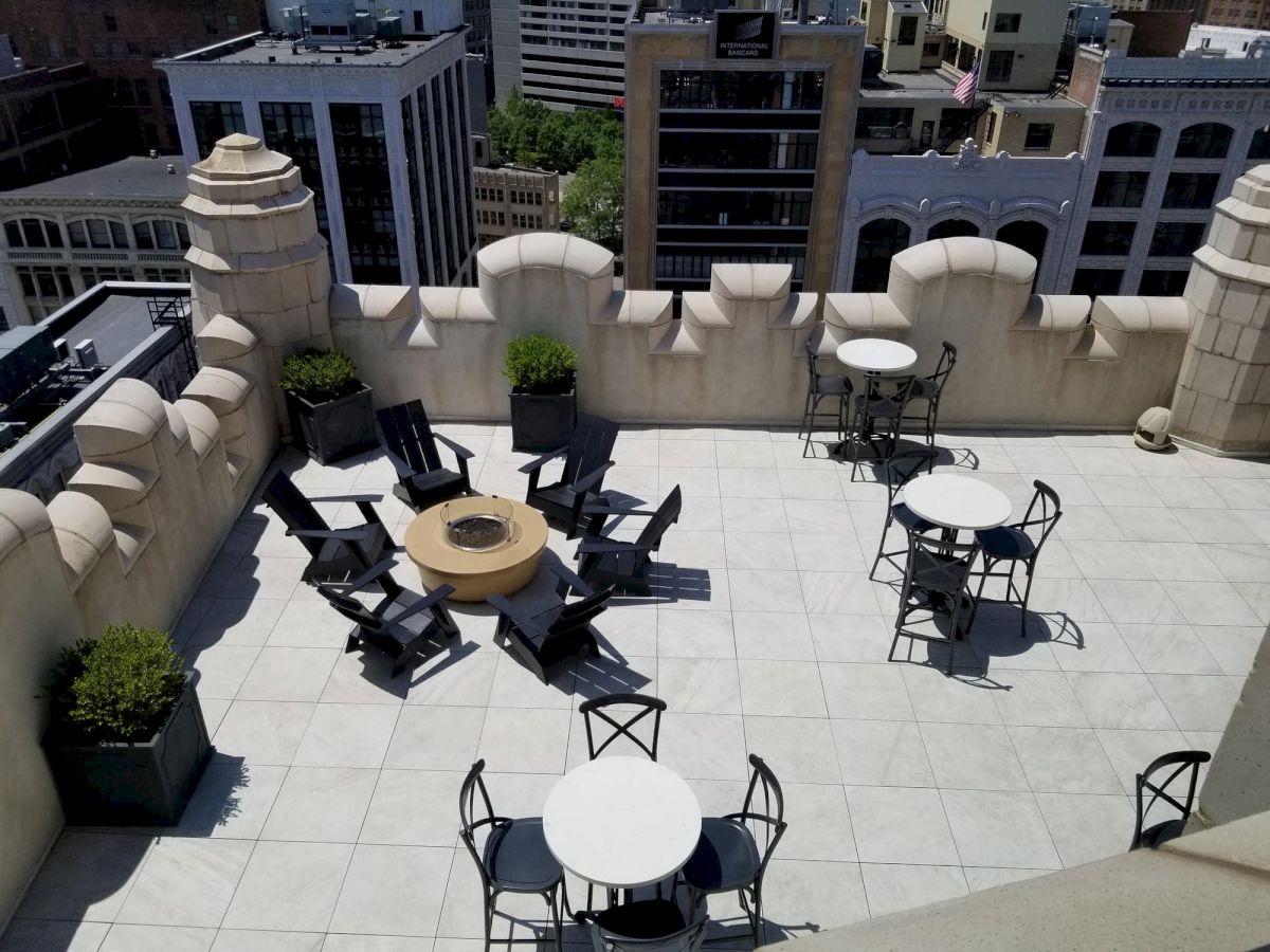 Rooftop terrace with a fire pit, chairs, and tables surrounded by a decorative wall with city buildings in the background.