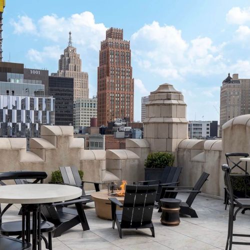 The image shows a rooftop patio with chairs, tables, and a fire pit, offering a view of city skyscrapers under a blue sky with clouds.