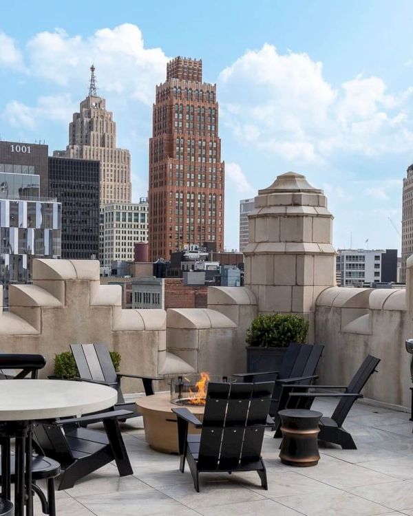 The image shows a rooftop patio with chairs, tables, and a fire pit, offering a view of city skyscrapers under a blue sky with clouds.