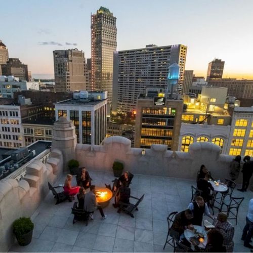 Rooftop gathering at dusk with people seated around tables and a fire pit, surrounded by city buildings and a sunset sky in the background.