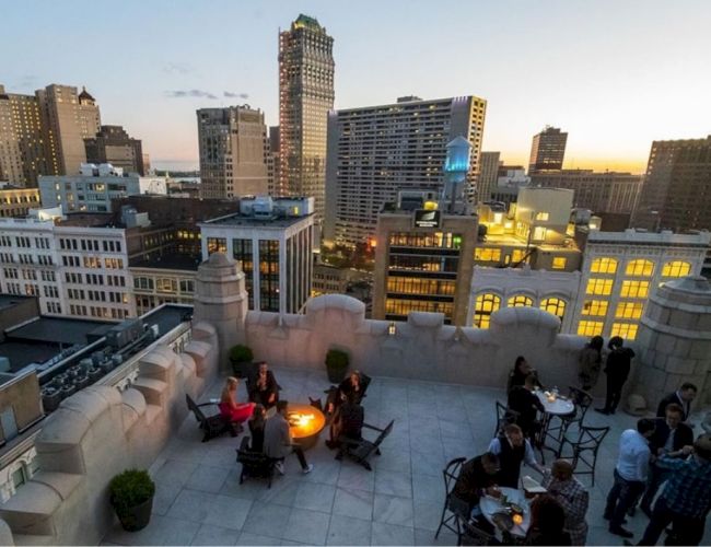 Rooftop gathering at dusk with people seated around tables and a fire pit, surrounded by city buildings and a sunset sky in the background.