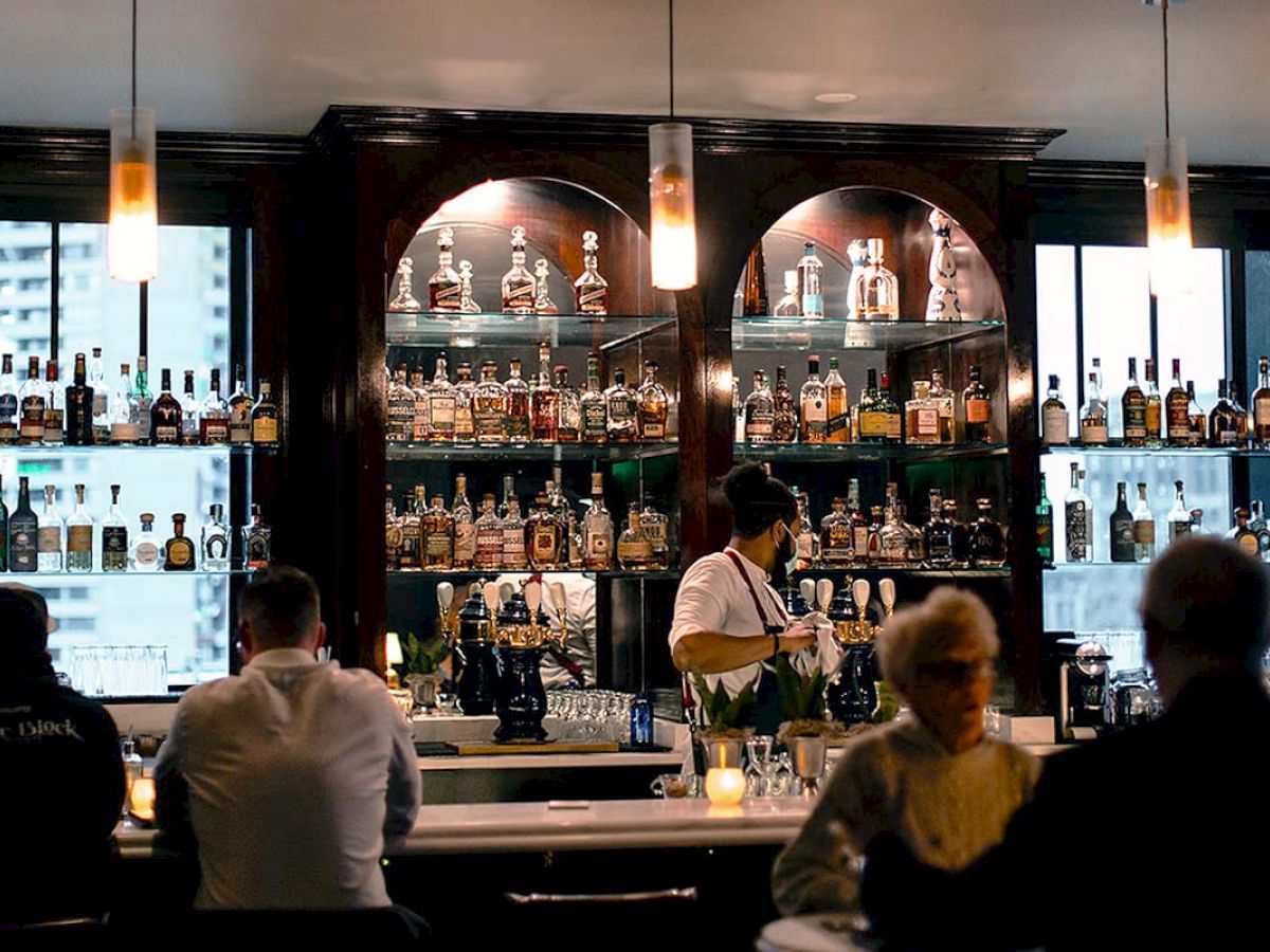 A dimly lit bar with a variety of bottles on shelves, a bartender serving drinks, and several patrons seated at the counter.