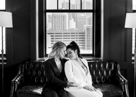 A black and white photo of two people sitting on a leather couch in front of large windows, embracing and smiling at each other.