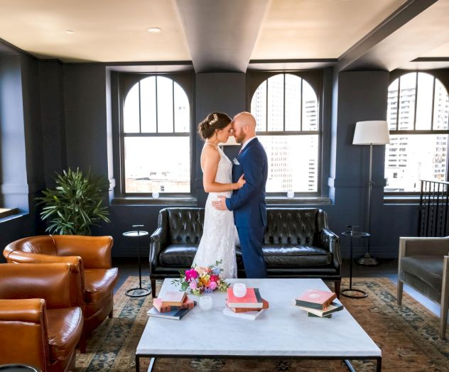 A couple shares a kiss in a stylish room with leather chairs, a coffee table with books and flowers, and large windows offering city views.