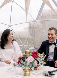 A couple in formal attire sits inside a decorative dome, with a colorful bouquet on the table in front of them and small trees nearby.