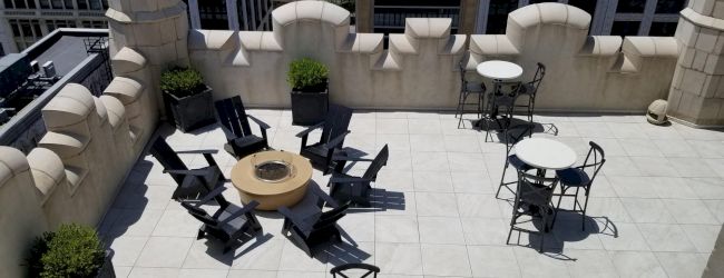 A rooftop terrace with tables, chairs, and a fire pit is shown, surrounded by decorative walls and urban buildings in the background.
