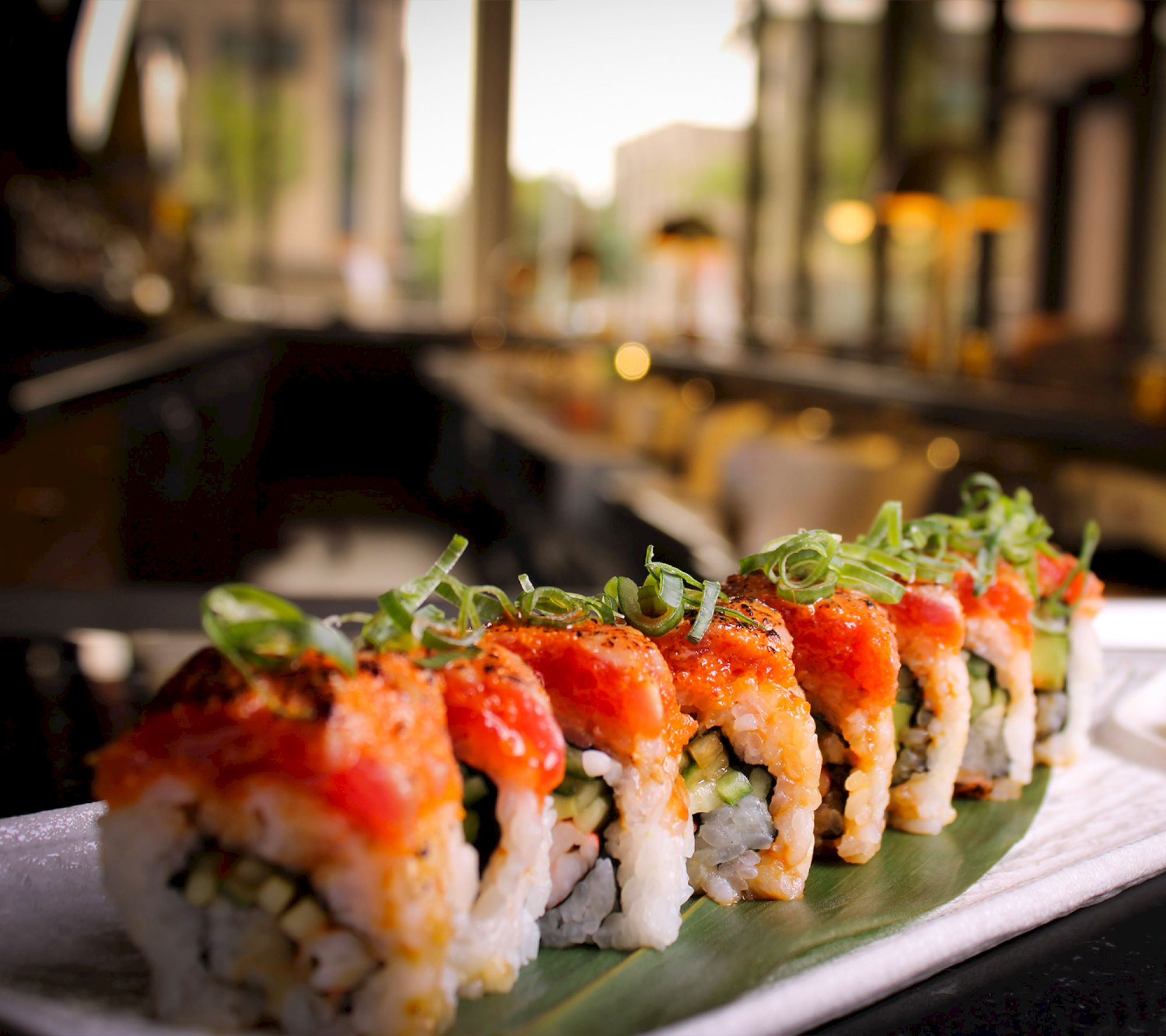 A line of sushi rolls topped with vibrant red seafood and green onion slices on a decorative leaf, set in a dimly lit restaurant.
