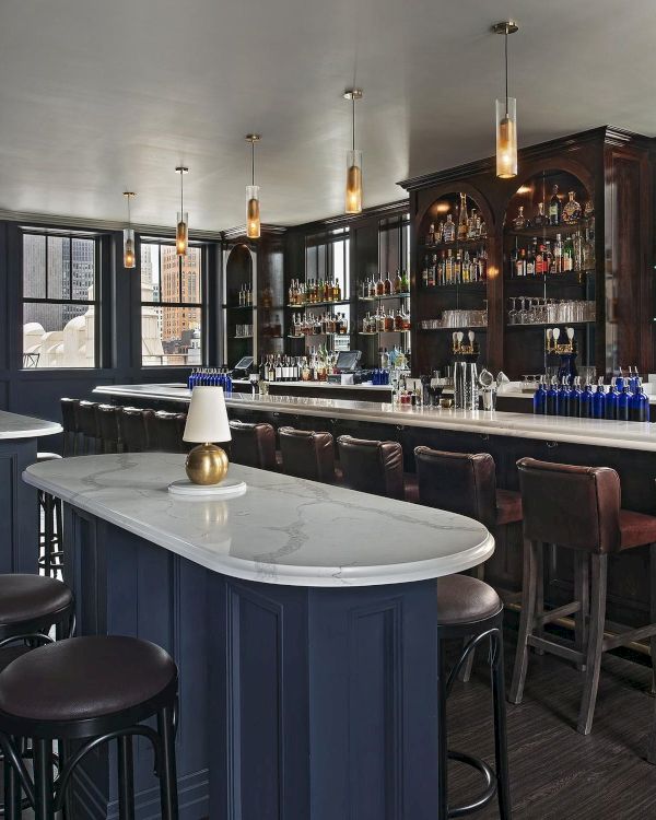 The image shows a cozy bar interior with a long counter, bar stools, shelves of bottles, and elegant pendant lighting providing ambience.