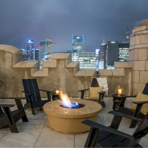 A rooftop patio features a fire pit surrounded by chairs, with city skyscrapers visible in the background under a night sky.