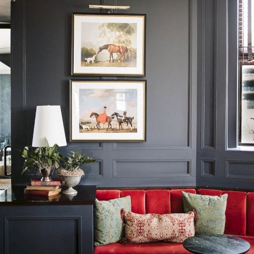 A stylish room with dark walls, red velvet seating, framed horse art, a small table, and decorative plants and books on the side.