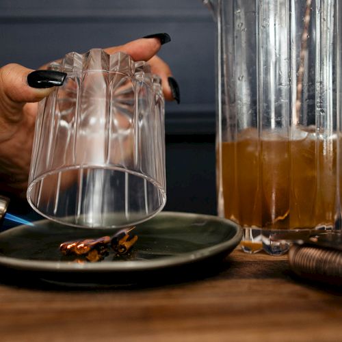 A hand holds a glass over a torched ingredient on a plate, next to a pitcher with a brown drink, possibly part of a cocktail preparation.