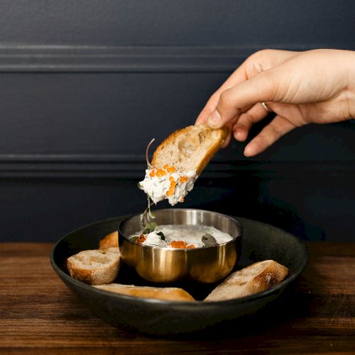 A hand dips a piece of bread into a bowl of creamy dip on a wooden table, surrounded by more bread in a black dish.