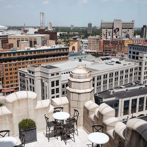Rooftop view of a cityscape with buildings, outdoor tables, chairs, and greenery. Signs and structures are visible in the background.