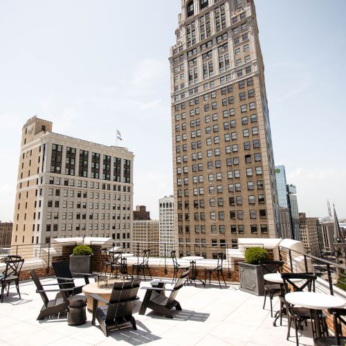 Rooftop patio with tables and chairs, surrounded by tall buildings under a clear sky.