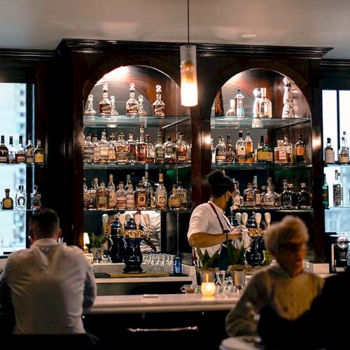 A bartender serves drinks in a dimly lit bar with patrons seated, surrounded by shelves of various liquor bottles.