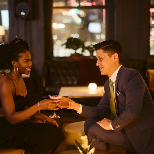 A couple sits in a dimly lit lounge, smiling and clinking glasses. The atmosphere is cozy, with warm lighting and leather chairs.