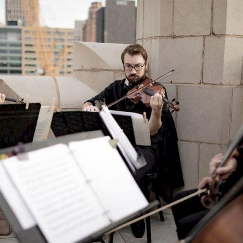 A string quartet is performing outdoors on a rooftop with music sheets in front of them, reflecting a cityscape backdrop.