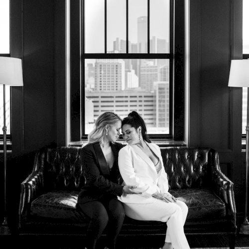 A black and white image of two individuals sitting closely on a sofa, in front of a window with a city skyline view.