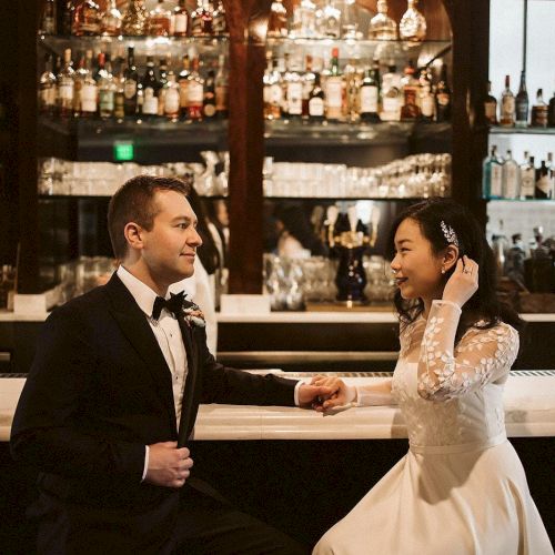 A couple in wedding attire sits at a bar, surrounded by bottles, engaging in conversation.