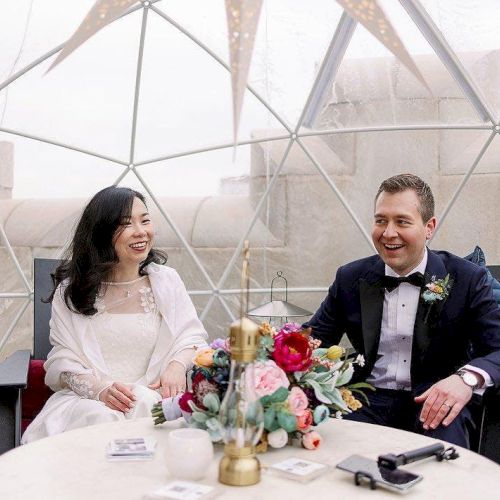 A couple is seated inside a dome-shaped structure, dressed formally, with flowers on the table in front of them.