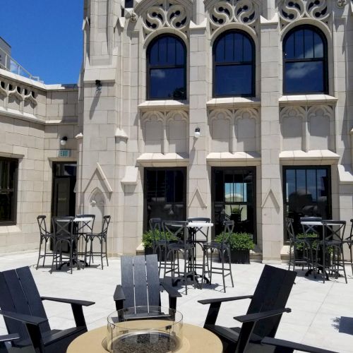 An outdoor patio with chairs and tables surrounds a fire pit in front of a building with ornate windows and detailed architecture.