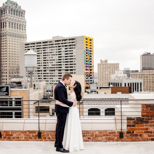 A couple embraces on a rooftop with a city skyline in the background, including tall buildings and colorful architectural details.
