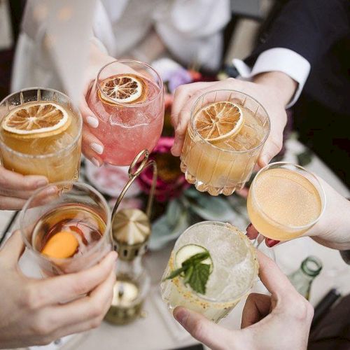 People are holding various colorful cocktails garnished with fruit slices, toasting together in a celebratory manner.