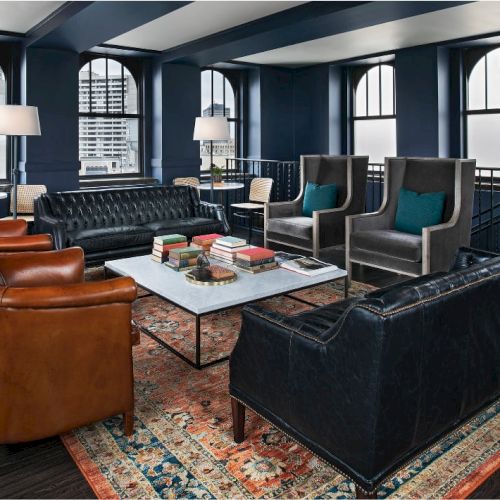 A stylish lounge with leather chairs, sofas, a coffee table, lamps, and large windows, featuring a patterned rug and a cityscape view.