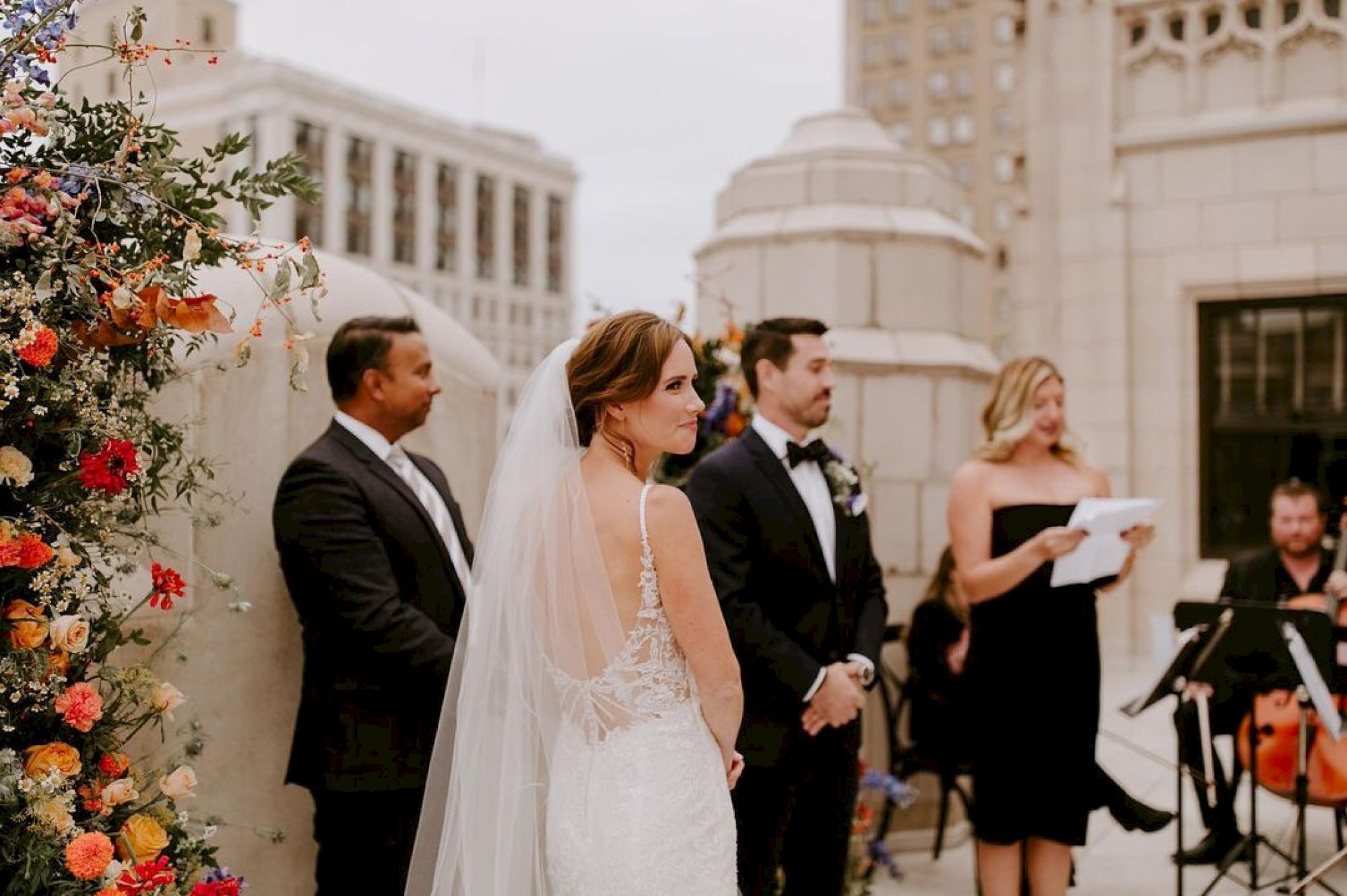 A wedding ceremony is taking place outdoors, with a bride, groom, and officiant present, alongside a musician and a floral arrangement.