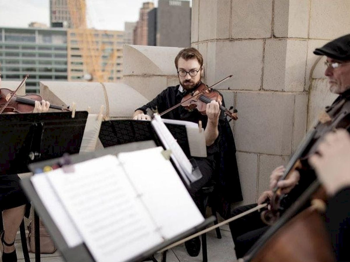 A string quartet performs on a rooftop, with musicians focused on their sheet music and instruments, against a cityscape backdrop.